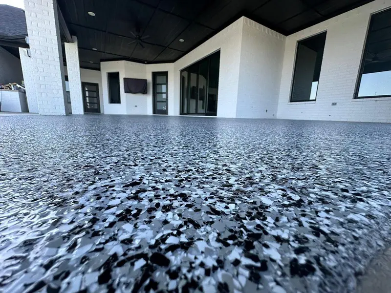 Low-angle close-up of a gray, white, and black flake patio coating beside a modern home